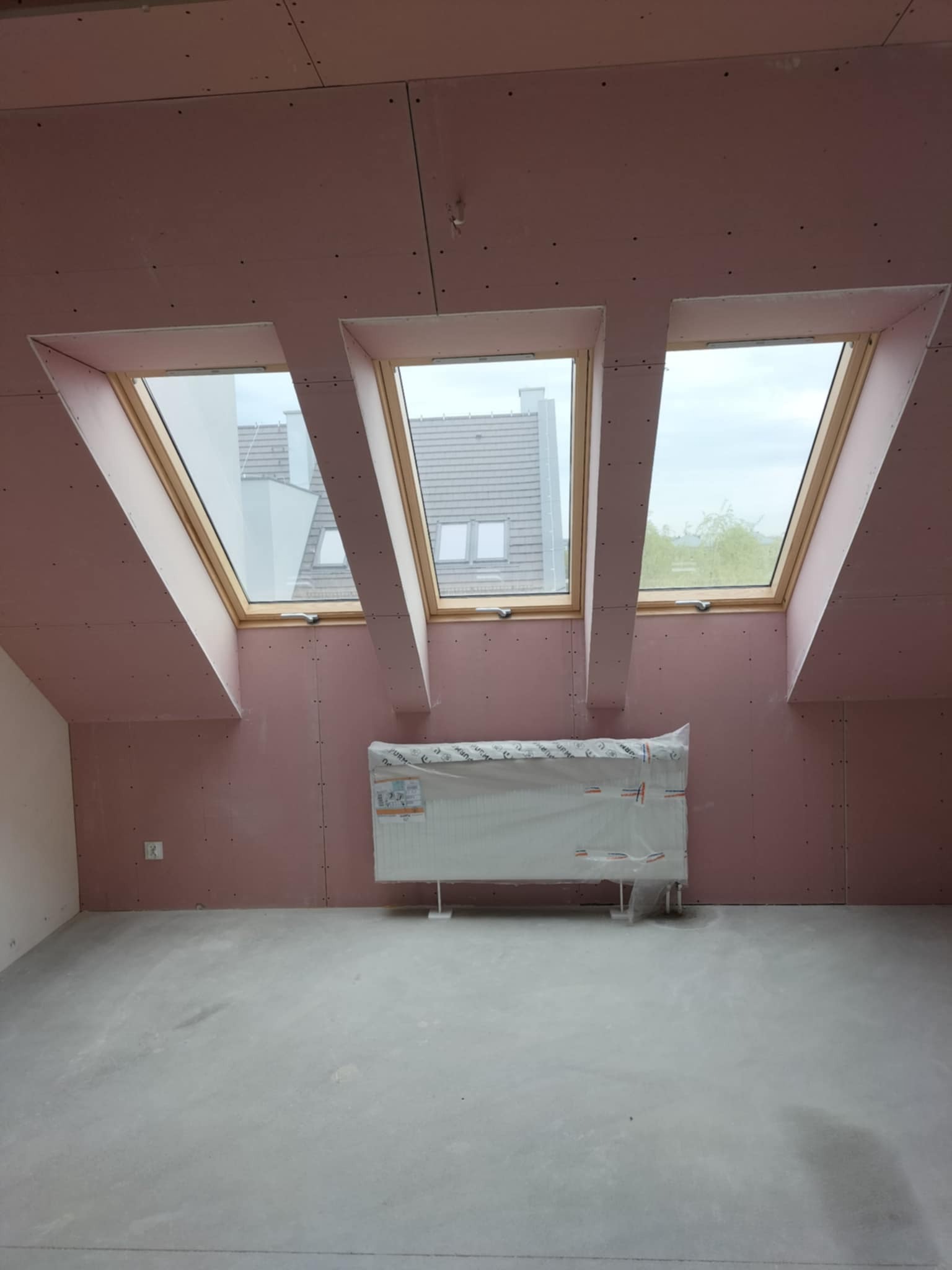 Poddasze z trzema oknami dachowymi, ściany wyłożone płytami karton-gips w kolorze różowym, grzejnik zabezpieczony folią, surowa podłoga.
