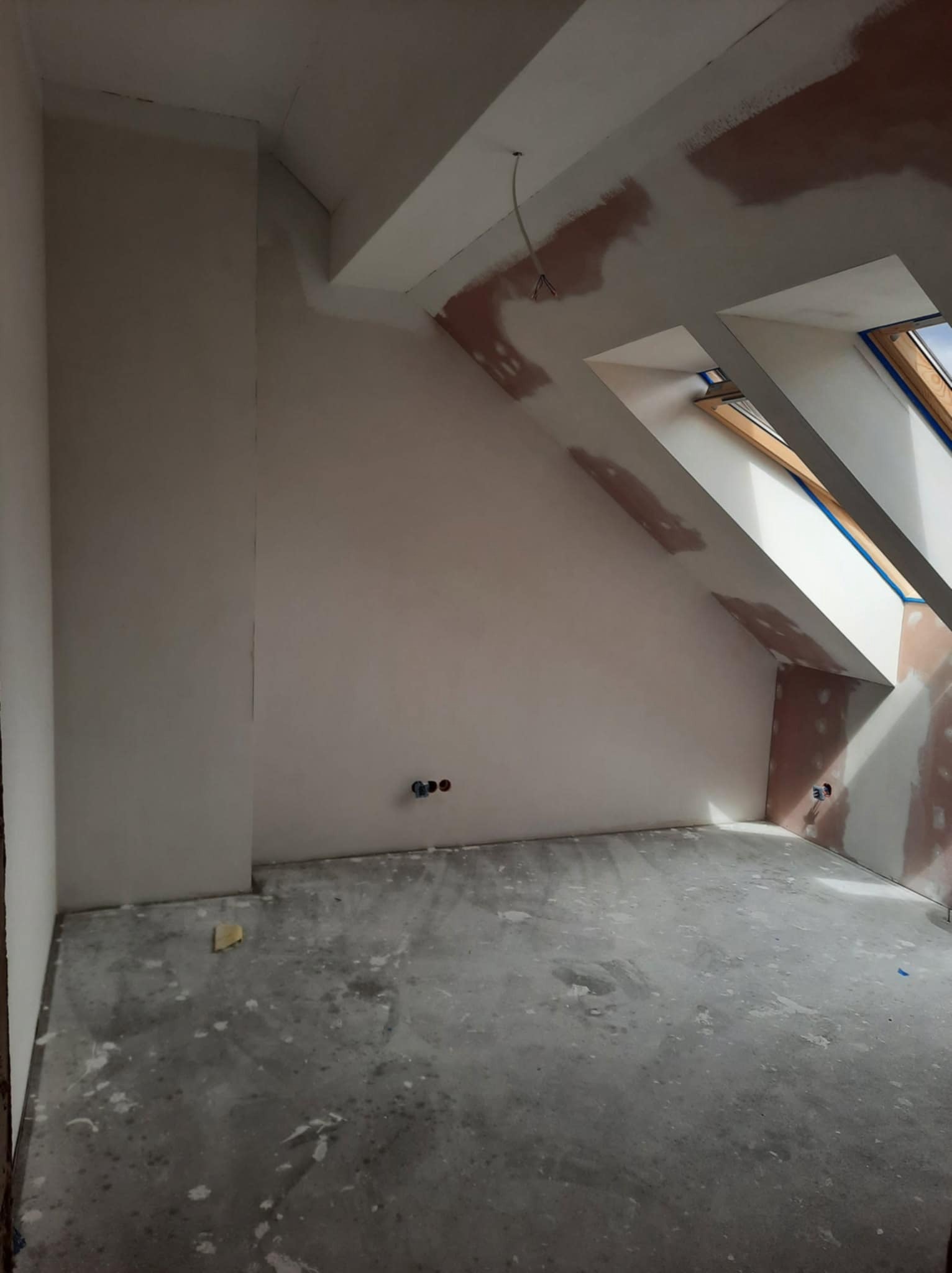 Pokój na poddaszu w trakcie prac wykończeniowych: ściany przygotowane pod gładź, widoczne okna dachowe i wystające kable. Podłoga zabezpieczona przed zabrudzeniem.