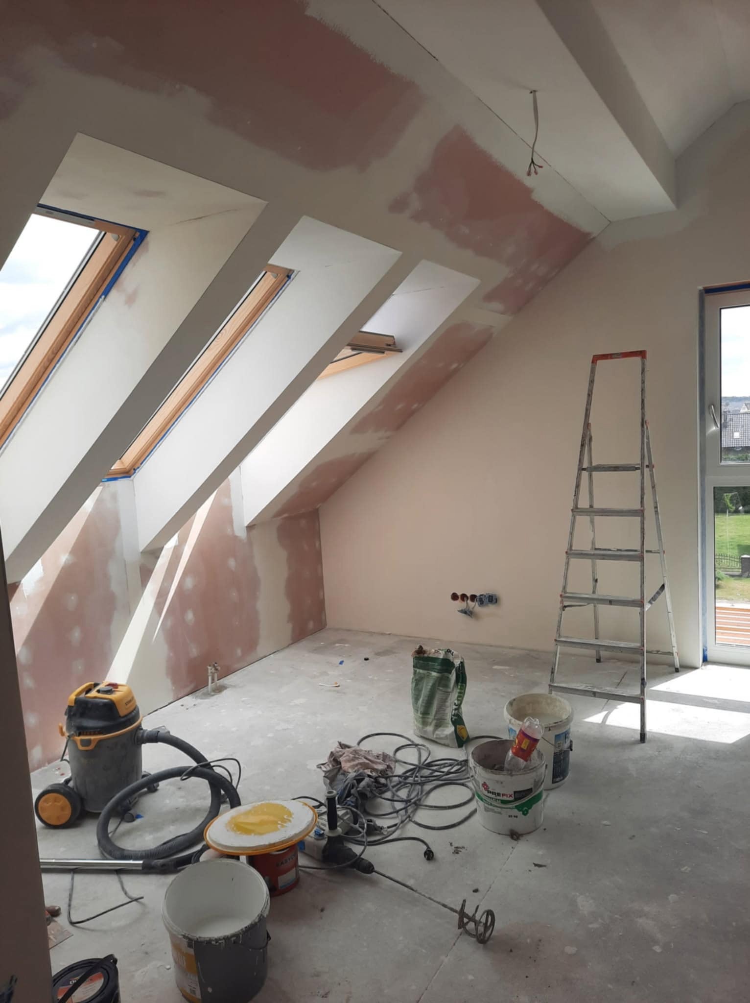 Poddasze w trakcie gładzi, widoczne okna dachowe, drabina, narzędzia i materiały budowlane. Ściany przygotowane pod malowanie.