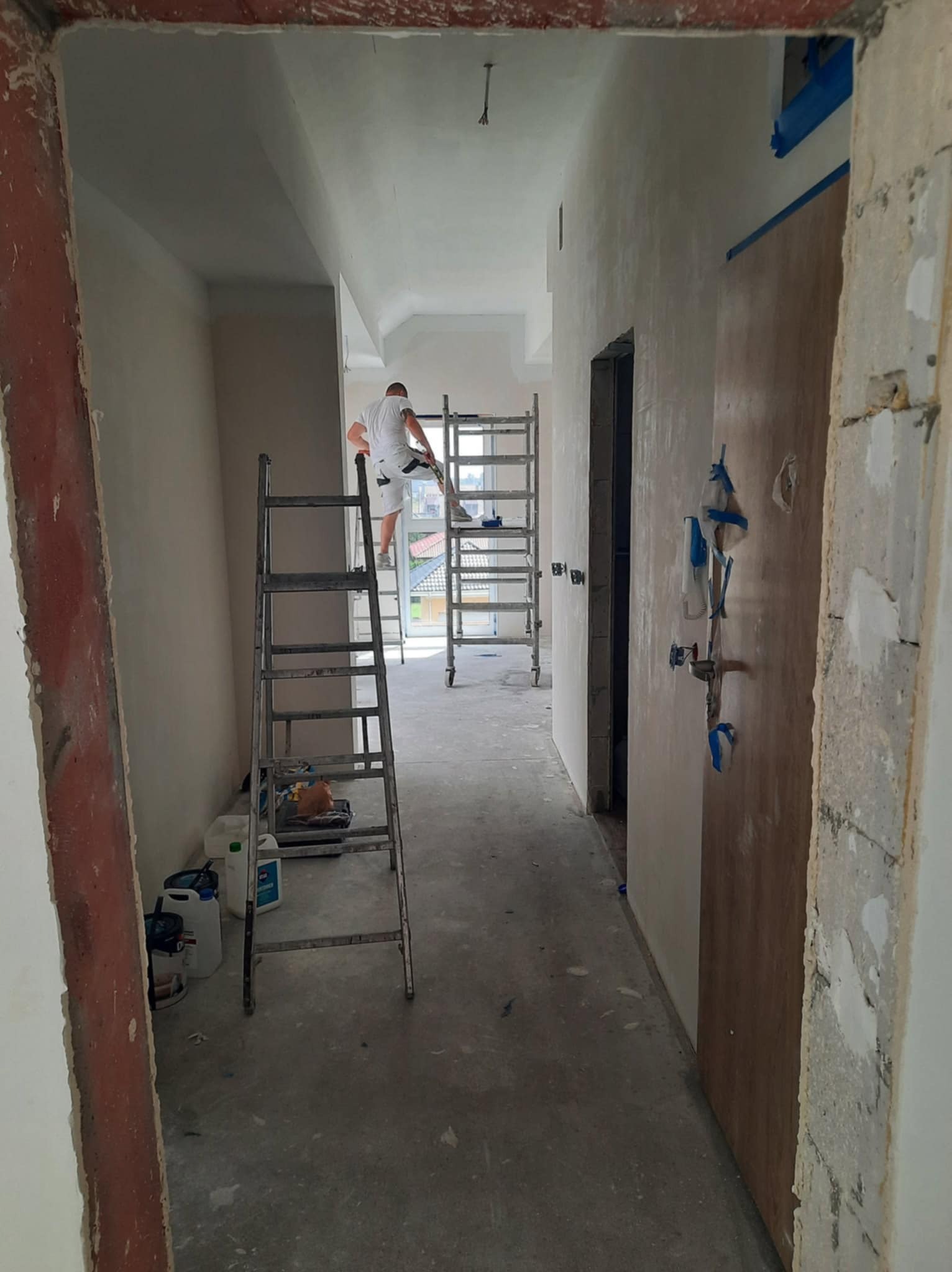 Remont korytarza: mężczyzna szpachluje ścianę na rusztowaniu, widoczne drabiny, wiaderka z farbą i zabezpieczone drzwi. Korytarz w trakcie prac wykończeniowych.