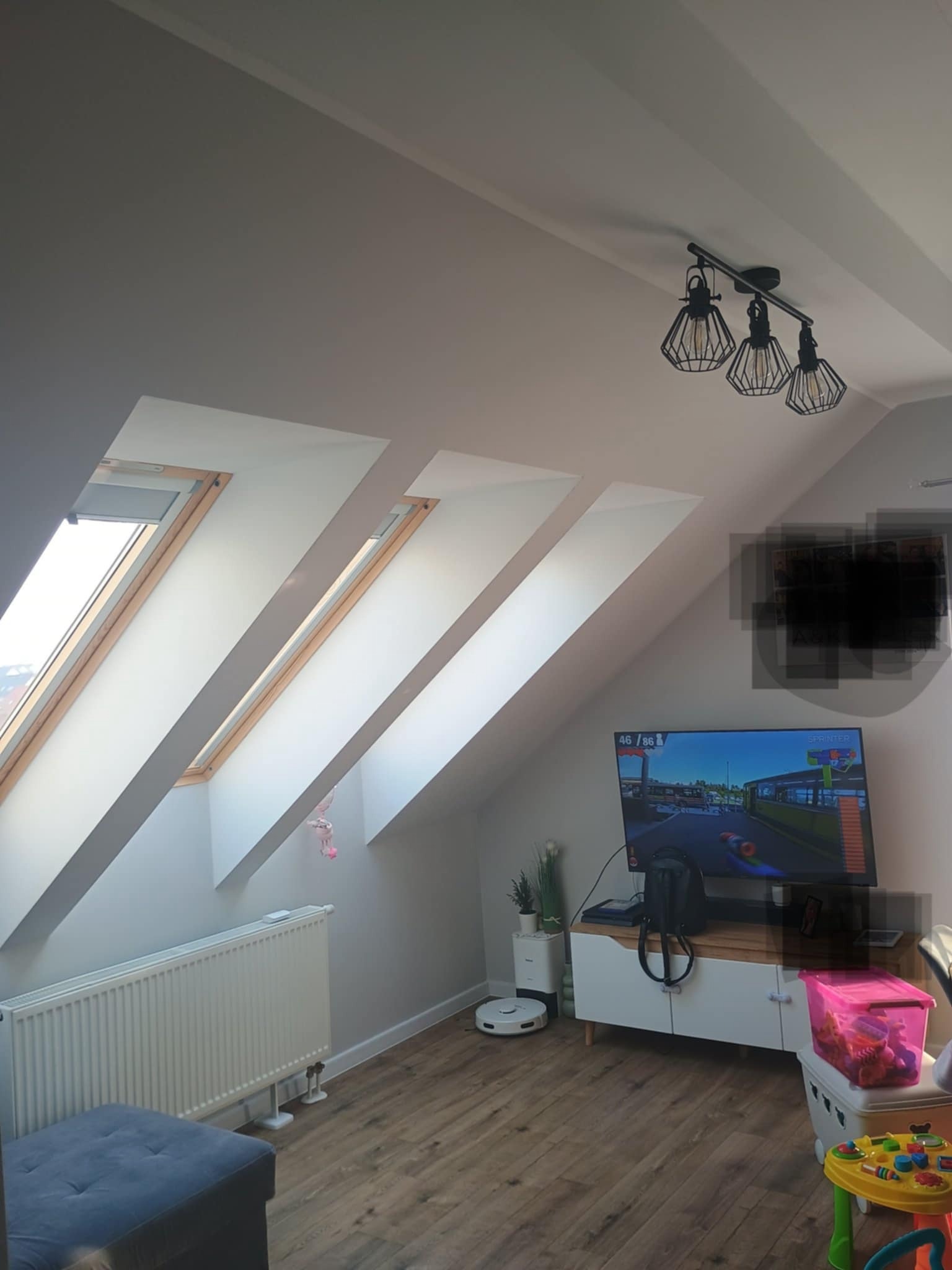 Pokój na poddaszu z trzema oknami dachowymi, grzejnikiem, telewizorem i drewnianą podłogą. Widoczna nowoczesna lampa sufitowa.