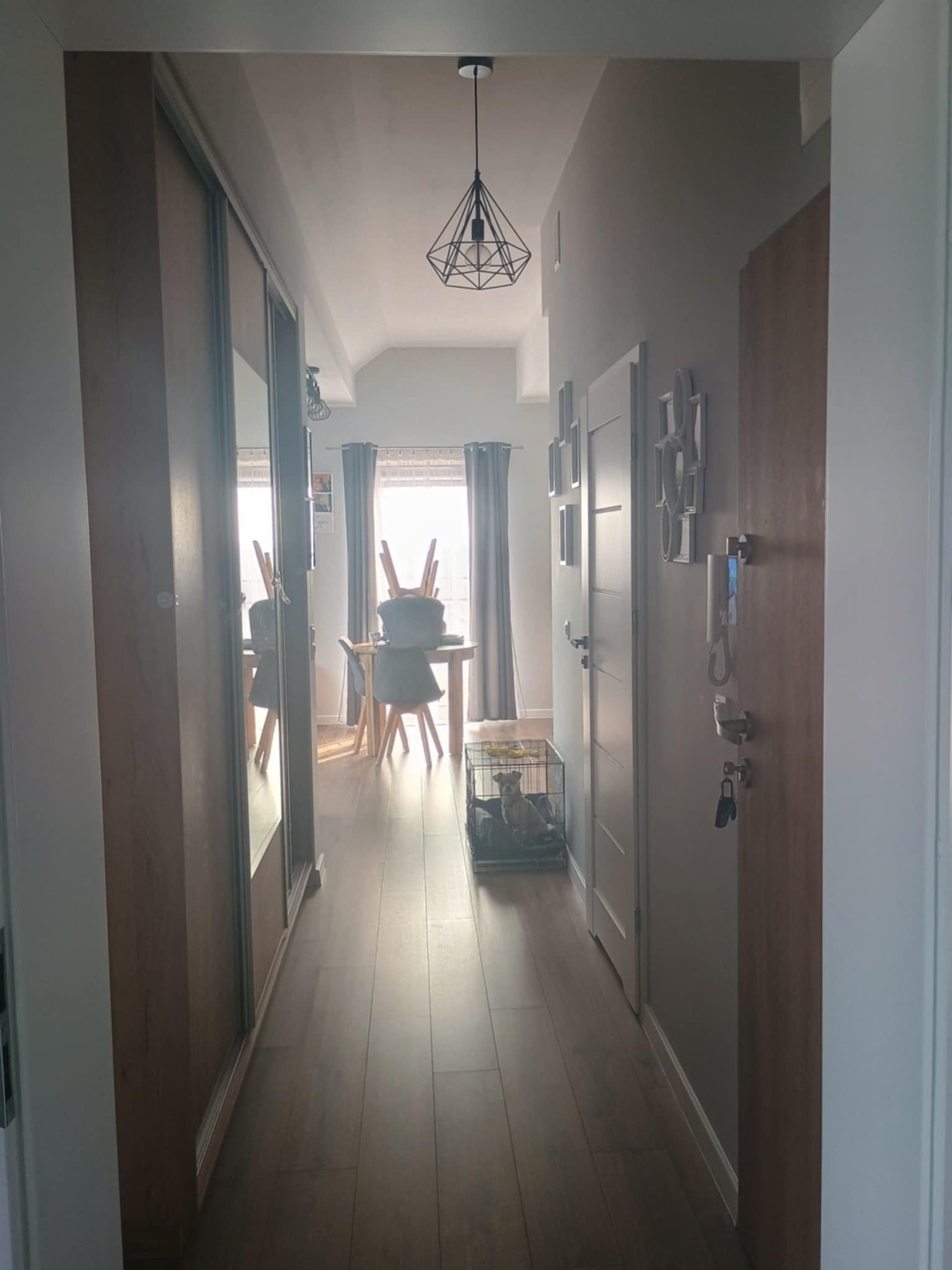 Nowoczesny korytarz z drewnianą podłogą, przesuwną szafą z lustrem i widokiem na jadalnię z geometryczną lampą. W tle okno.