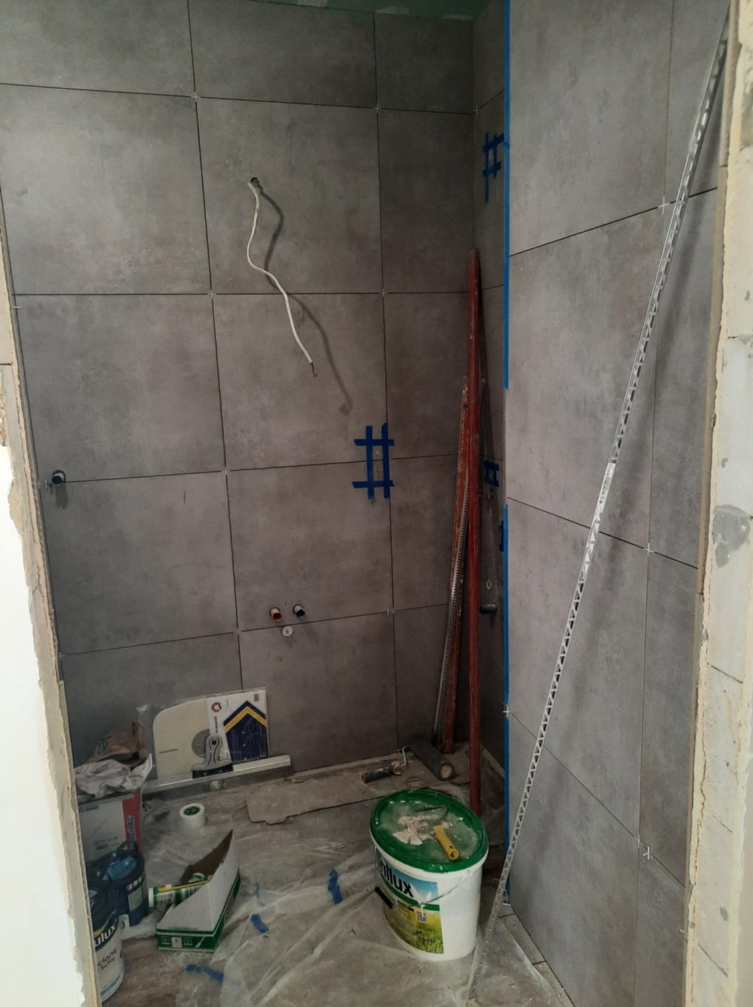 Szare płytki ścienne w trakcie układania z widocznymi krzyżykami dystansowymi, puszkami elektrycznymi i narzędziami. Fragment instalacji elektrycznej i wiadro z farbą.