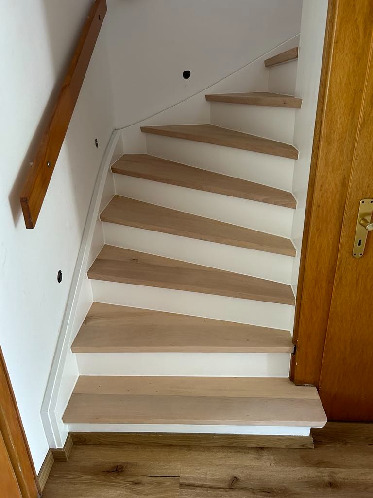 Drewniane schody z białymi podstopnicami i drewnianą poręczą. Widoczne wykończenie podłogi z paneli. Ujęcie od dołu schodów.