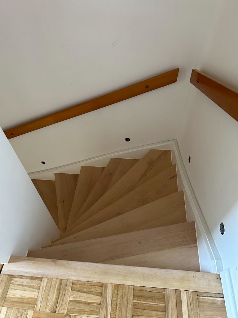 Drewniane schody spiralne z parkietem w jodełkę, widok z góry. Naturalne oświetlenie podkreśla strukturę drewna. Listwy przypodłogowe i białe ściany.