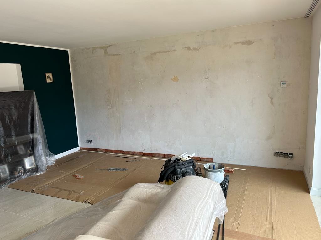 Przygotowanie ścian do gładzi: zabezpieczona podłoga, surowe ściany z widocznymi śladami po naprawach, zielona ściana w tle. Widoczne narzędzia i materiały malarskie.