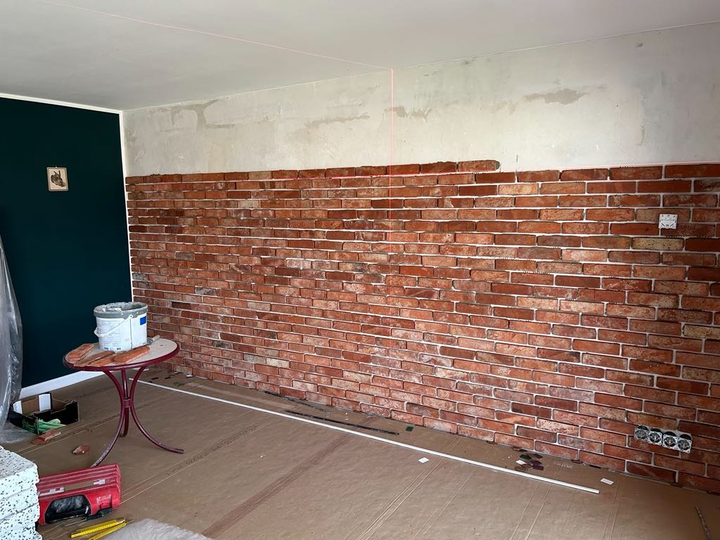 Ceglana ściana w trakcie montażu z widocznymi narzędziami i poziomnicą laserową. Częściowo zakończony montaż na tle surowej ściany i przygotowanego podłoża.