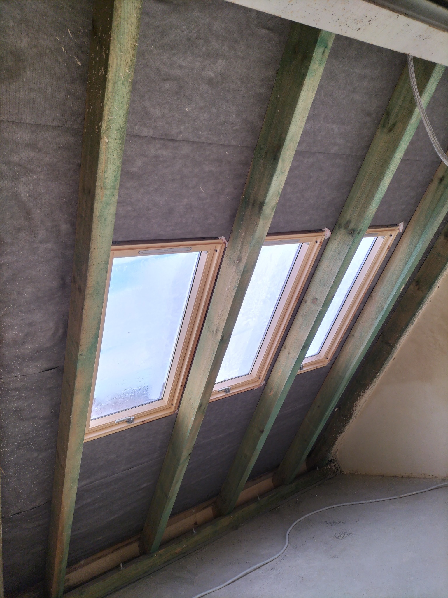 Montaż trzech okien dachowych w drewnianej konstrukcji dachu, widok od wewnątrz. Szara membrana dachowa i surowy beton na podłodze. Widoczne przewody elektryczne.