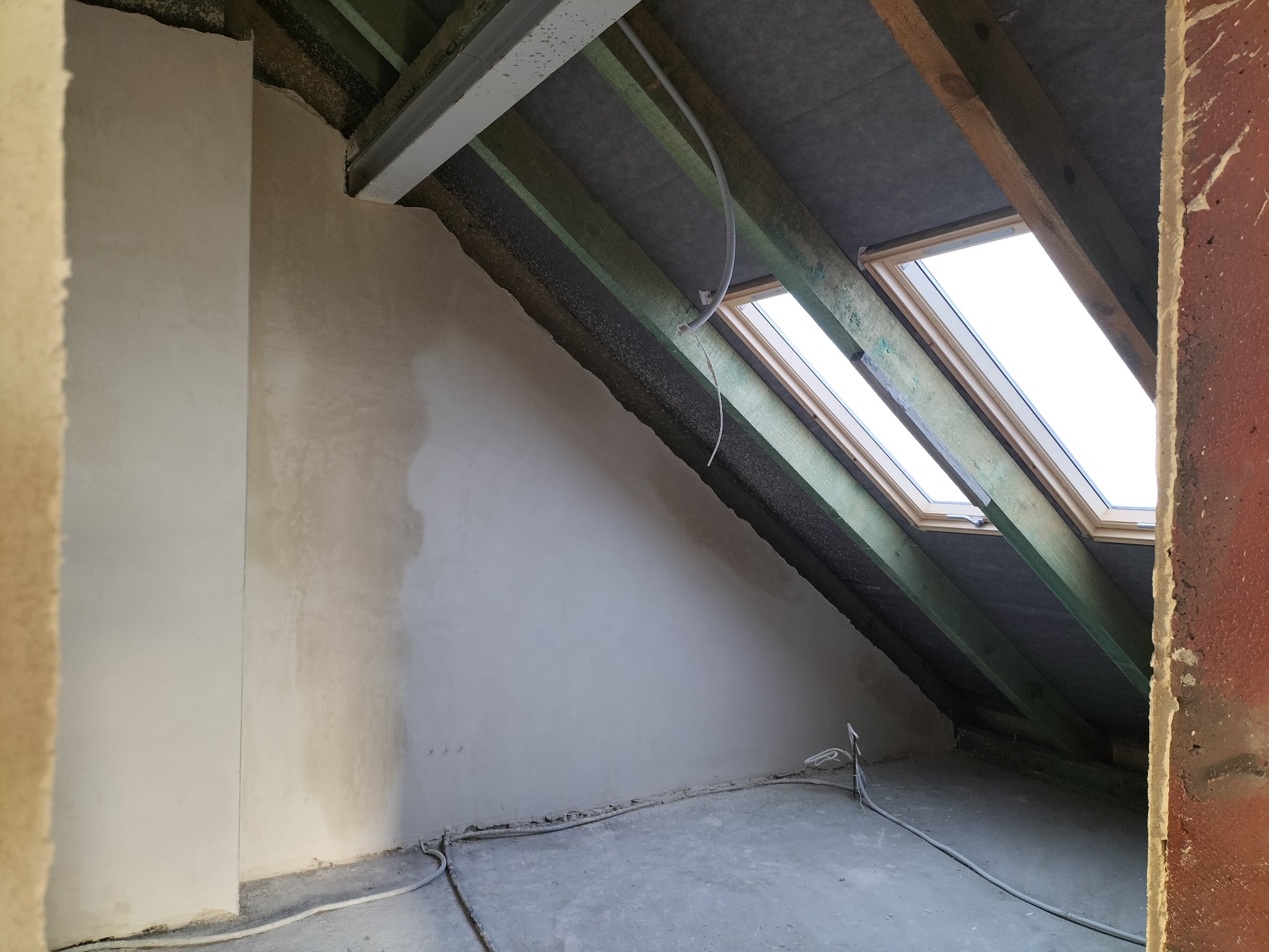 Poddasze w trakcie wykończenia: ściany po gładzi, widoczne okna dachowe, drewniana konstrukcja dachu i kable elektryczne przy podłodze. Surowy stan, przygotowanie do dalszych prac.