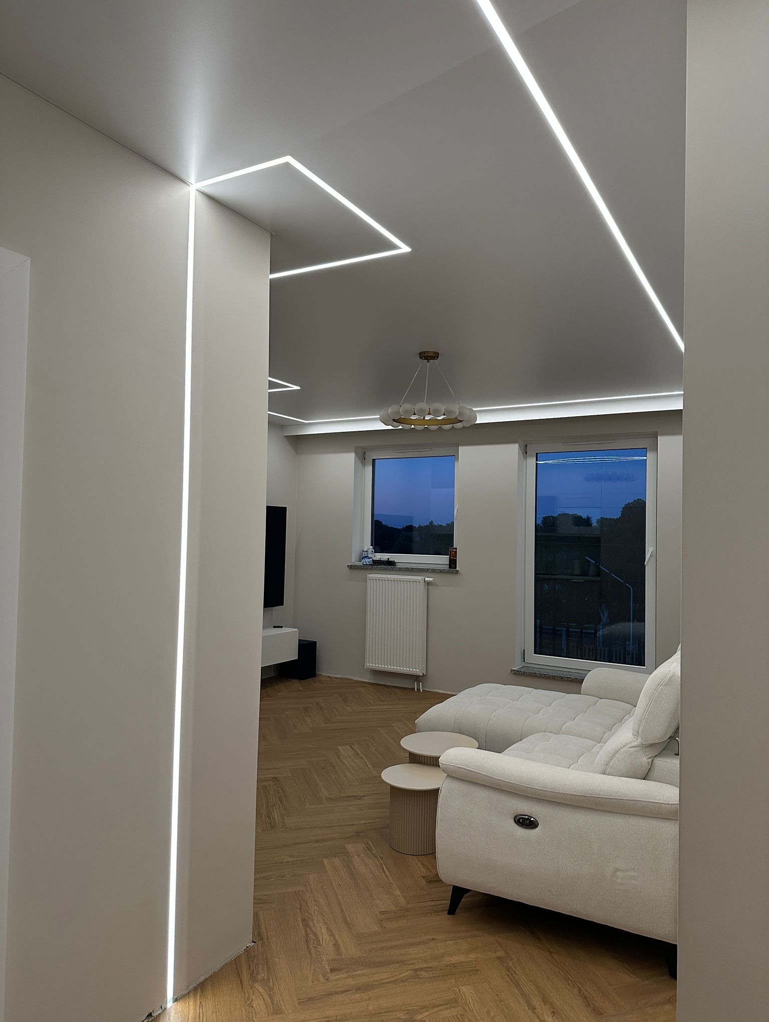 Nowoczesne oświetlenie LED w salonie: pionowe i poziome linie światła na ścianach i suficie, parkiet w jodełkę, biała sofa i widok na zewnątrz przez okno.