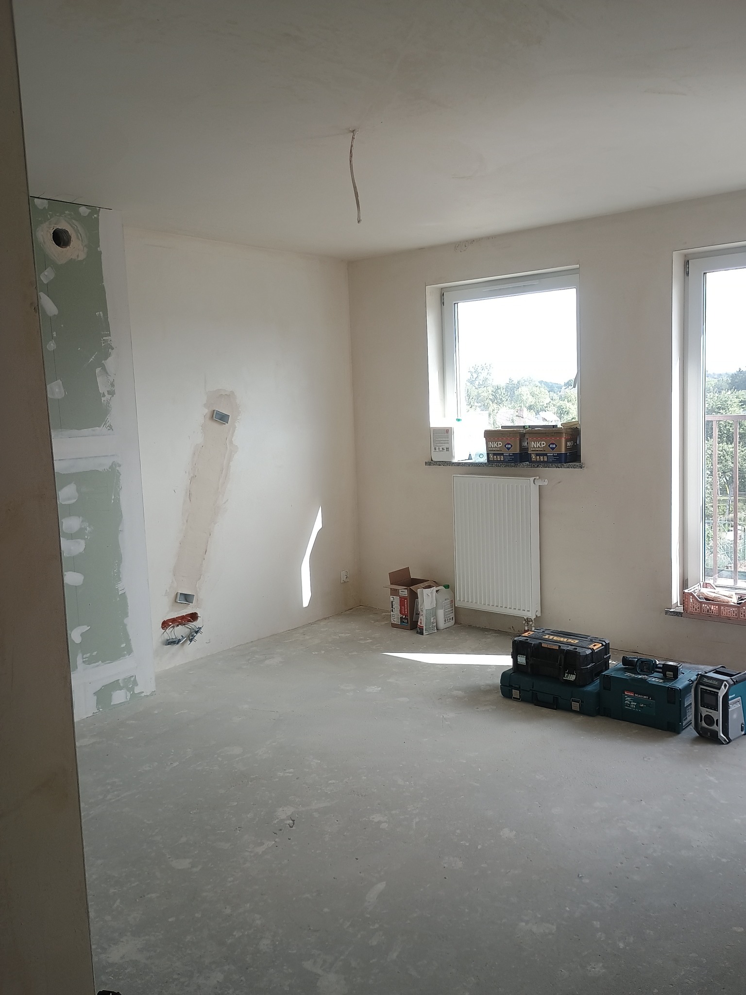 Pokój w trakcie remontu: ściany z gładzią, płyta karton-gips, narzędzia na podłodze, okna z widokiem na zieleń. Widoczne ślady prac instalacyjnych.
