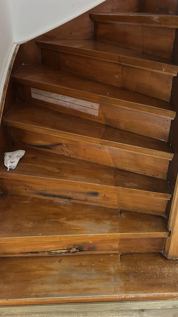 Drewniane schody w trakcie renowacji, widoczne ślady użytkowania i fragmenty starej powłoki. Na jednym ze stopni leży szmatka. Ujęcie z góry, naturalne światło.