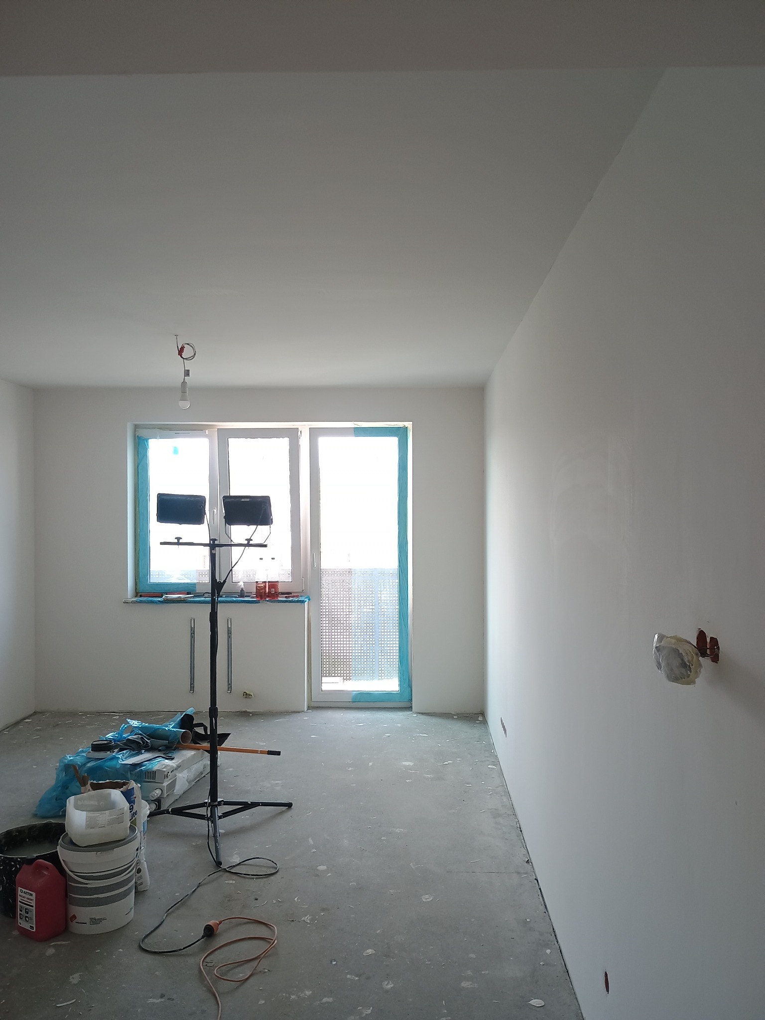 Wnętrze po gładziowaniu: białe ściany, okno zabezpieczone folią, sprzęt malarski na podłodze. Widoczne ślady farby na podłodze. Oświetlenie budowlane.