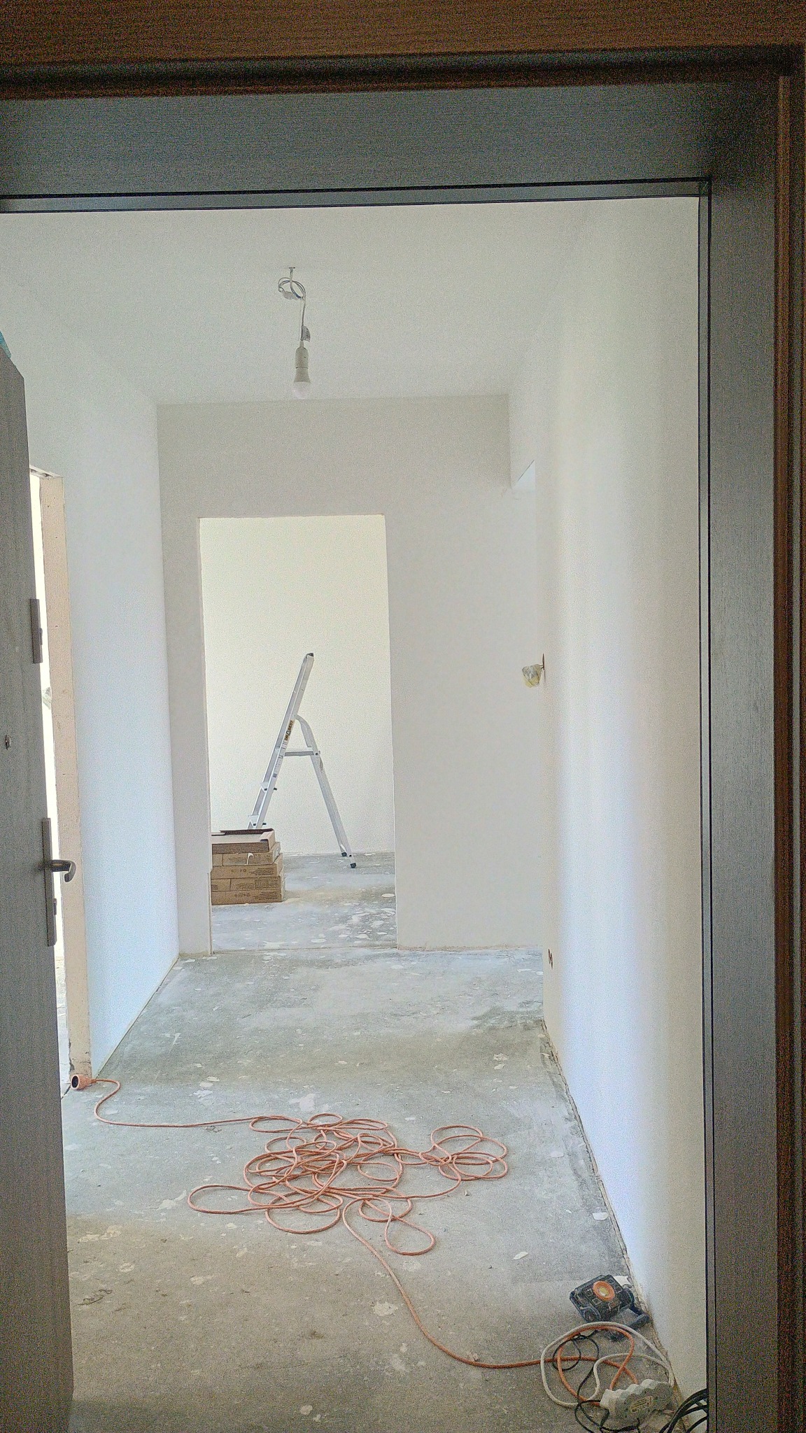 Wnętrze w trakcie remontu: białe ściany, surowa podłoga, drabina oparta o ścianę w głębi, zwój kabla na podłodze. Widok zza futryny drzwi.