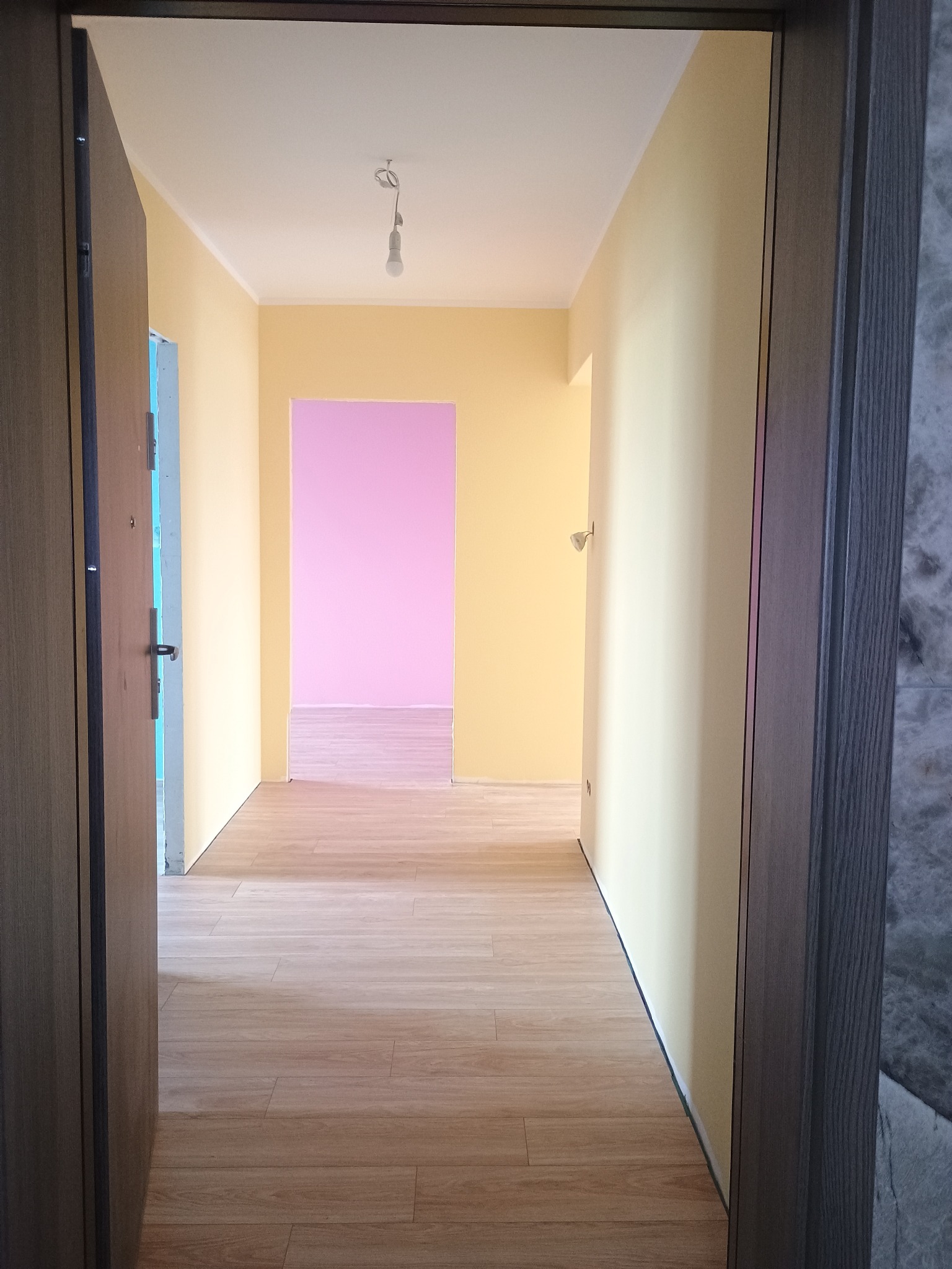 Wykończony korytarz z panelami podłogowymi i świeżo pomalowanymi ścianami w odcieniach żółci i różu, widok zza otwartych drzwi wejściowych.