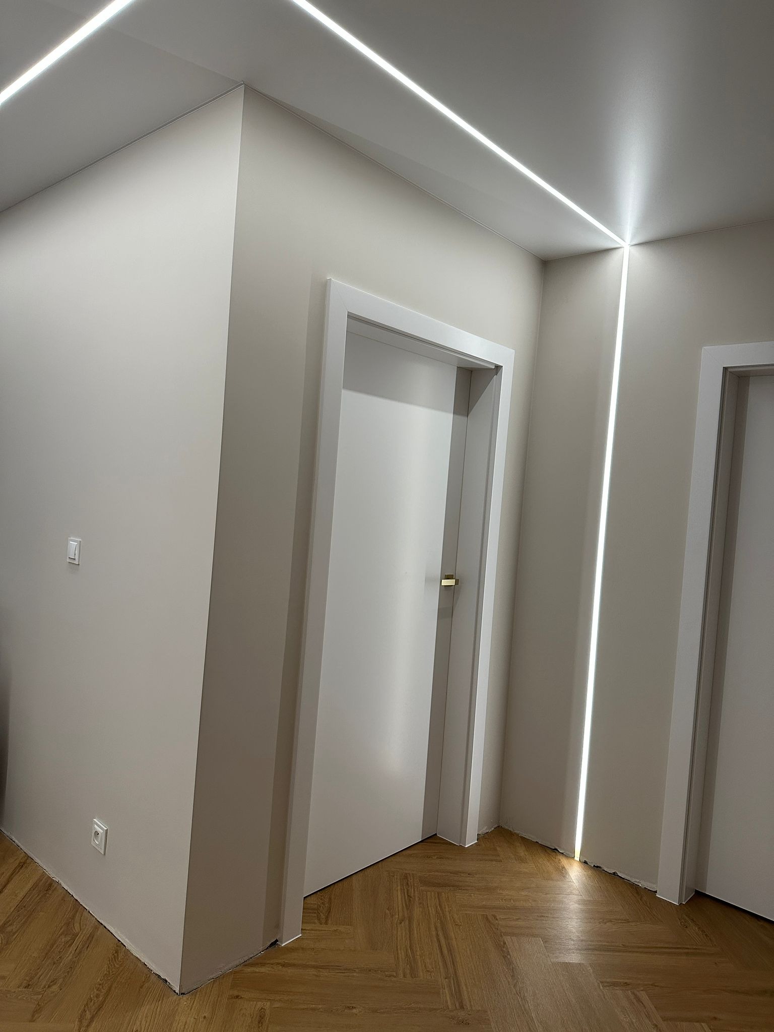 Nowoczesny korytarz z oświetleniem LED wzdłuż sufitu i ścian, białe drzwi, parkiet w jodełkę. Minimalistyczny design, jasne wnętrze, świeżo po remoncie.