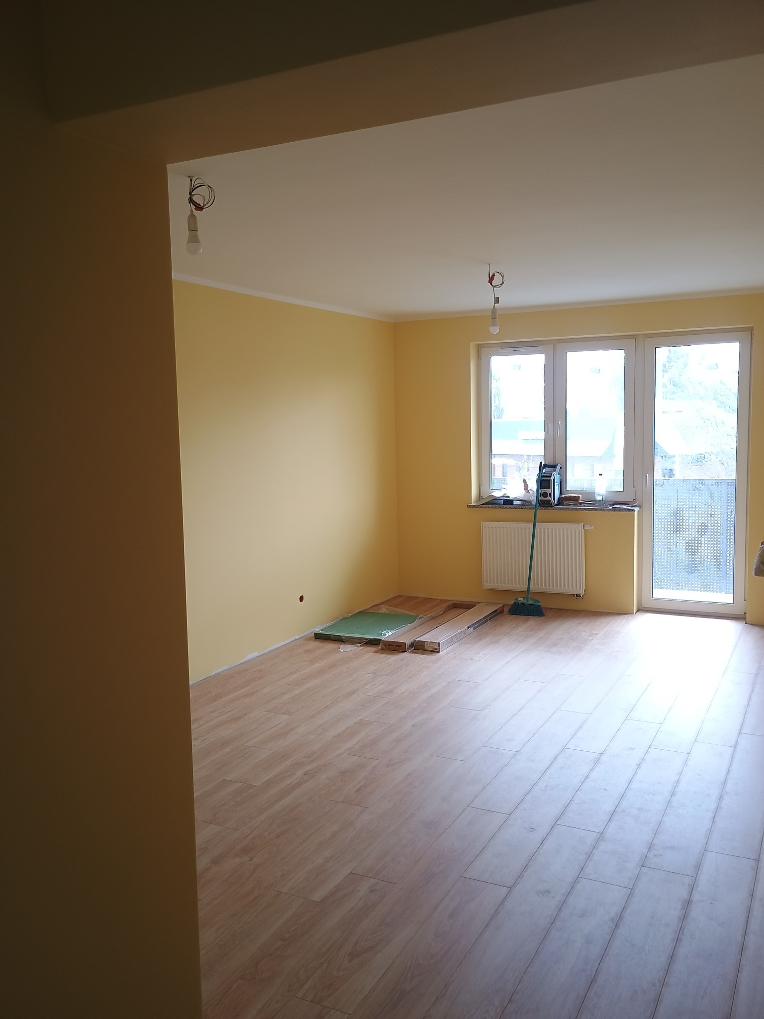 Jasne panele podłogowe w pokoju z żółtymi ścianami, widoczne kartony z panelami, okno balkonowe i grzejnik. Częściowo wykończone wnętrze.