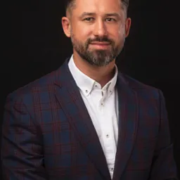 Portret mężczyzny z brodą w niebiesko-bordowej marynarce w kratę i białej koszuli na czarnym tle.