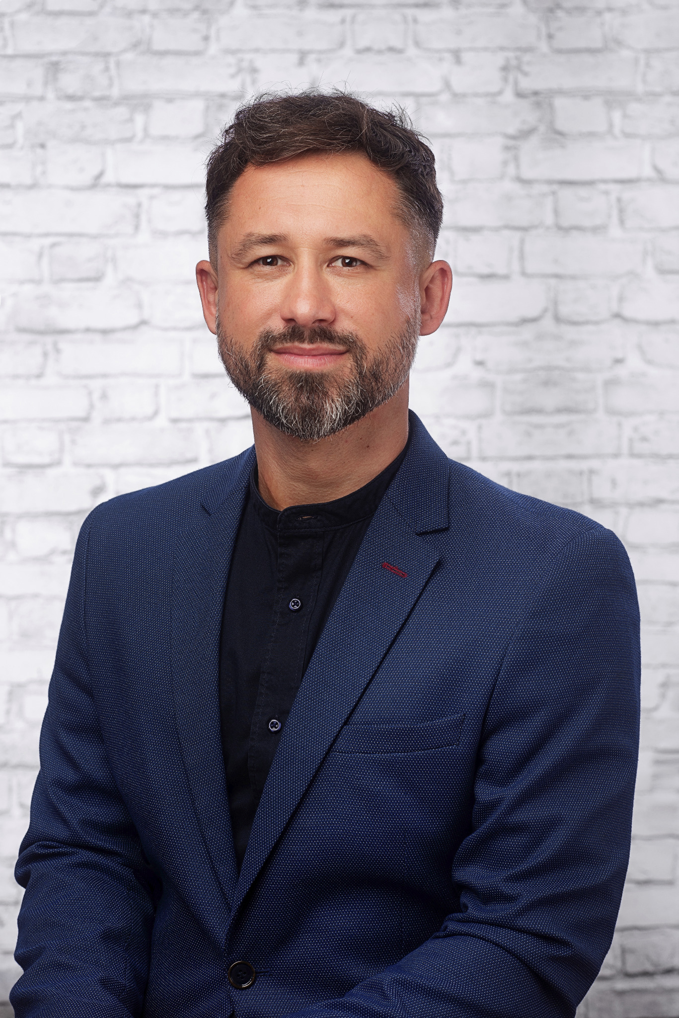 Portret mężczyzny z brodą w niebieskiej marynarce na tle białej ceglanej ściany.