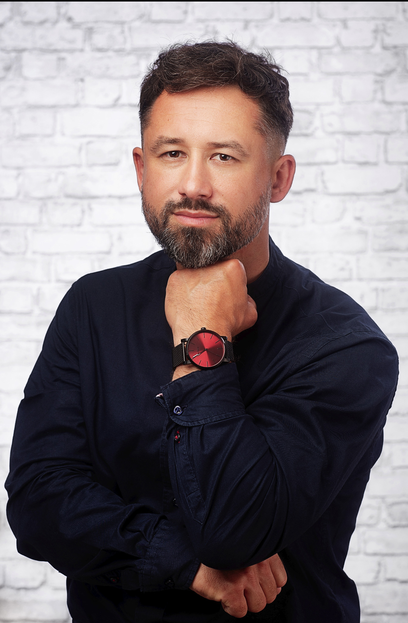 Portret mężczyzny z brodą, w ciemnej koszuli, z czerwonym zegarkiem na tle białej ściany z cegieł.