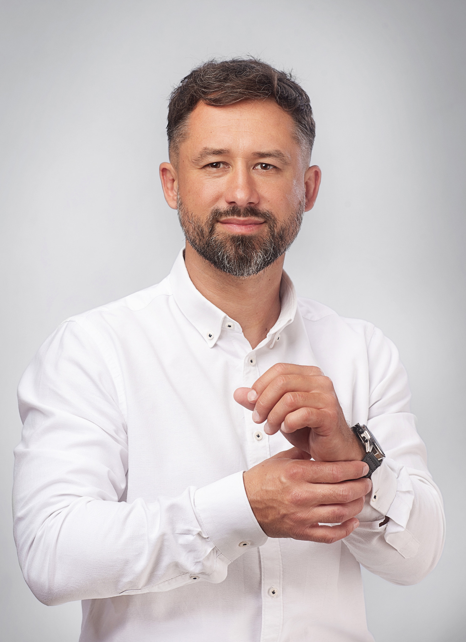 Portret mężczyzny w białej koszuli zapinającego mankiet zegarka na jasnym tle.