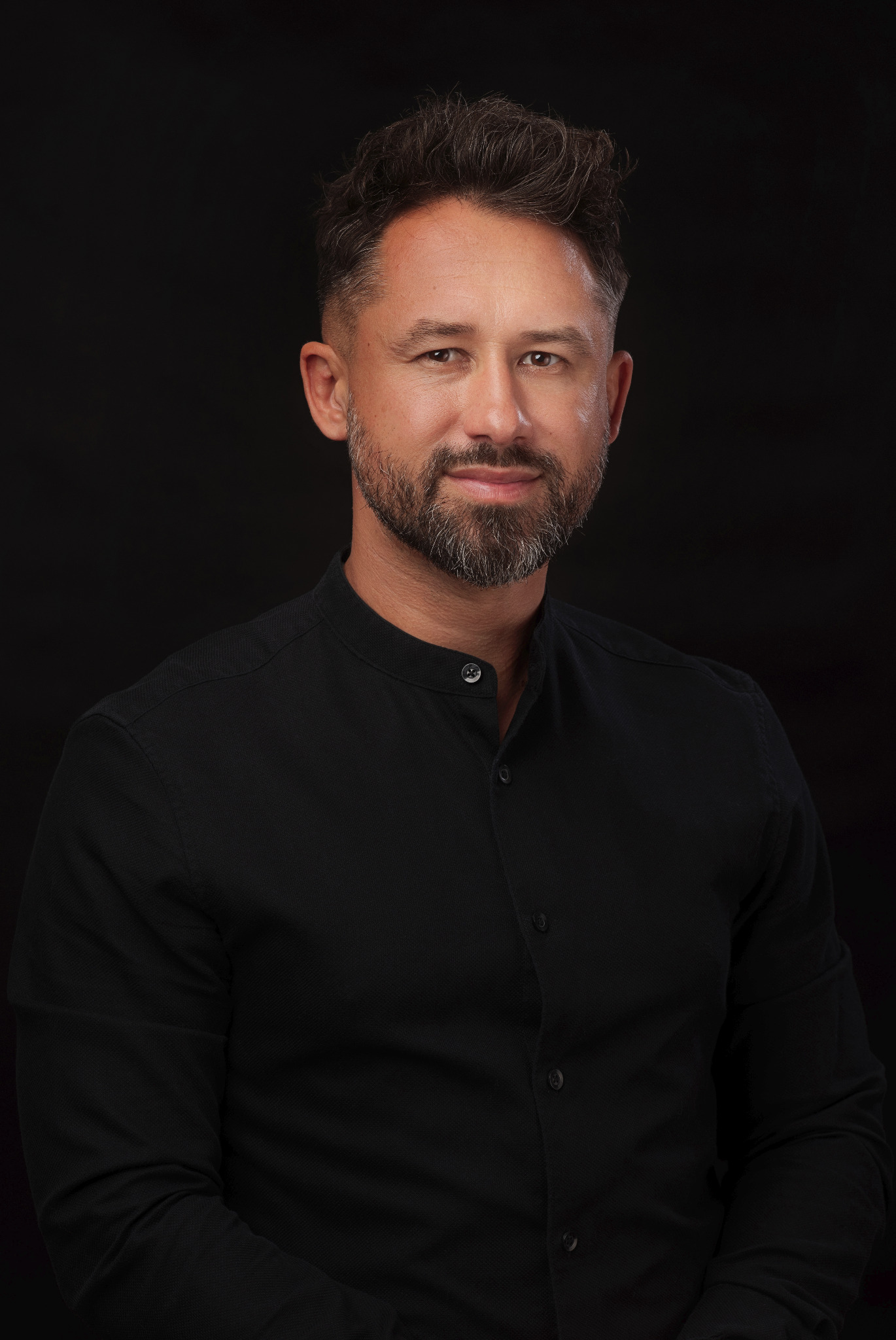 Portret mężczyzny z brodą w czarnej koszuli na ciemnym tle, lekki uśmiech, profesjonalne oświetlenie.