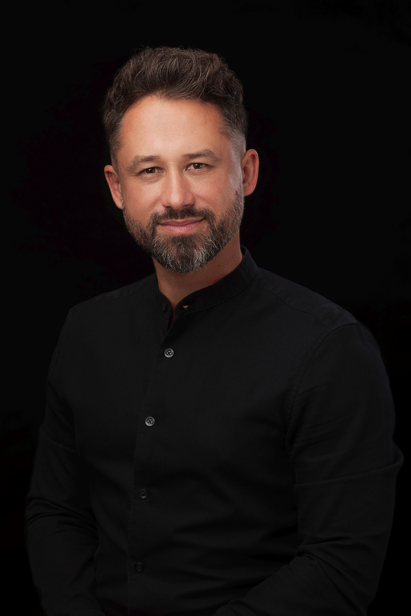 Portret mężczyzny z brodą w czarnej koszuli na ciemnym tle, uśmiechający się lekko.