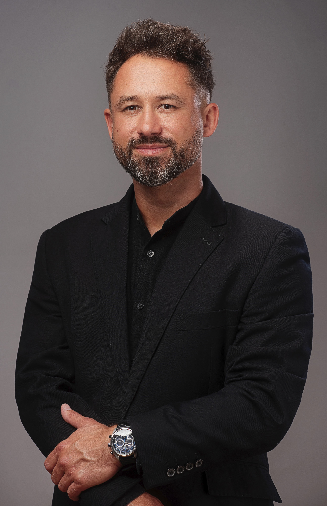 Portret mężczyzny w czarnym garniturze i koszuli, z brodą i zegarkiem na ręce, na szarym tle.