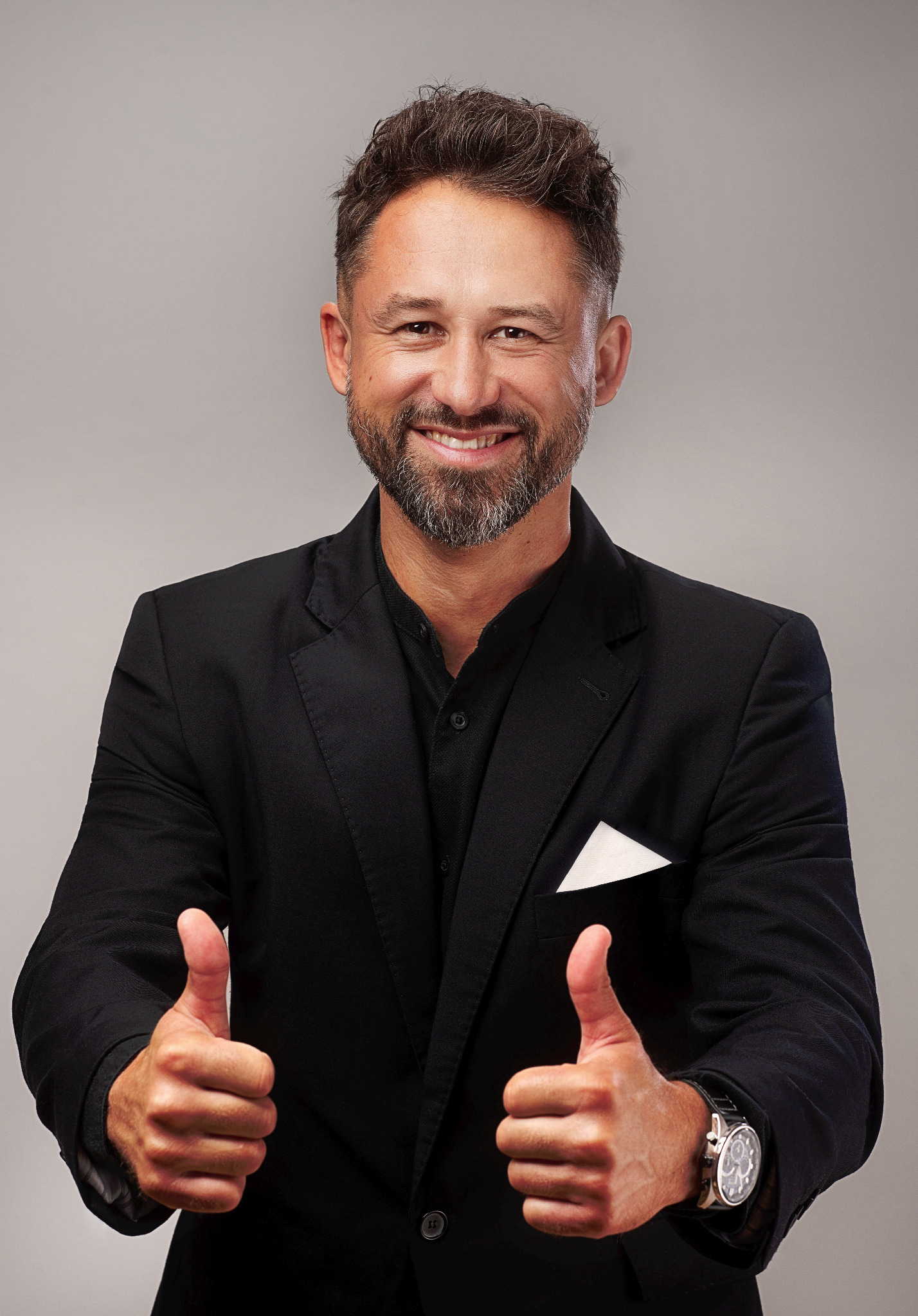 Uśmiechnięty mężczyzna w czarnym garniturze, z białą poszetką i zegarkiem, pokazuje dwa kciuki w górę na neutralnym tle.