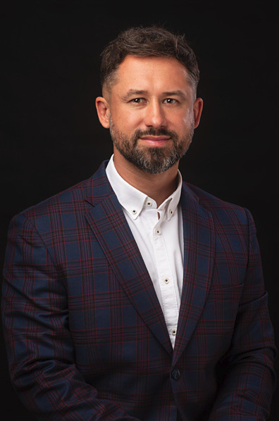 Portret mężczyzny z brodą w niebiesko-bordowej marynarce w kratę i białej koszuli na czarnym tle.