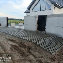 ART-KOP Artur Kwidziński - Ułożona ażurowa nawierzchnia z betonowych płyt na podjeździe przed nowoczesnym domem z betonowymi słupkami ogrodzenia i czarną bramą wjazdową, częściowo zanieczyszczona błotem.