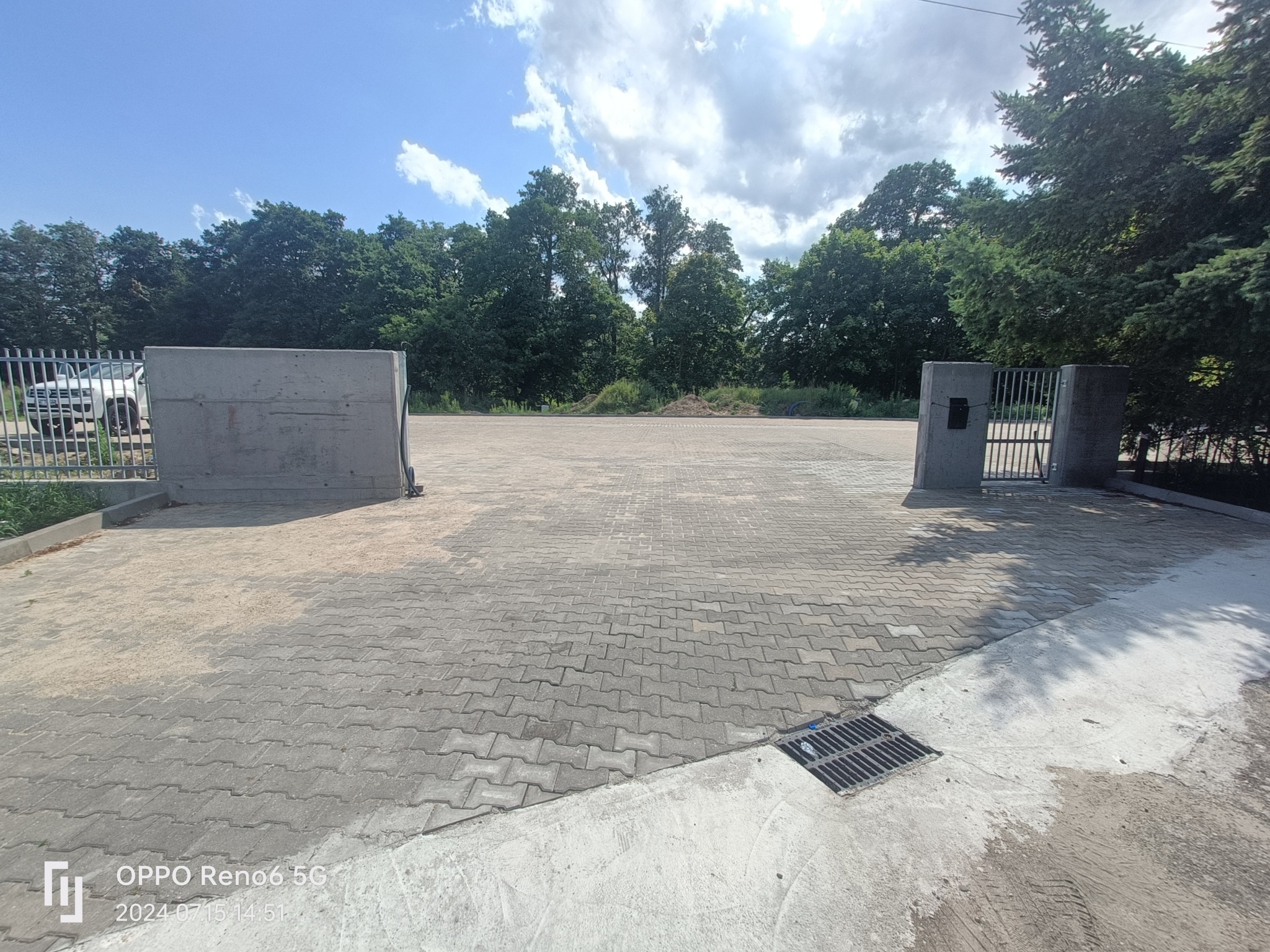 Rozległy plac wyłożony szarą kostką brukową, otoczony zielenią drzew i ogrodzeniem, z widocznym odpływem liniowym i fragmentem zaparkowanego pojazdu w tle.