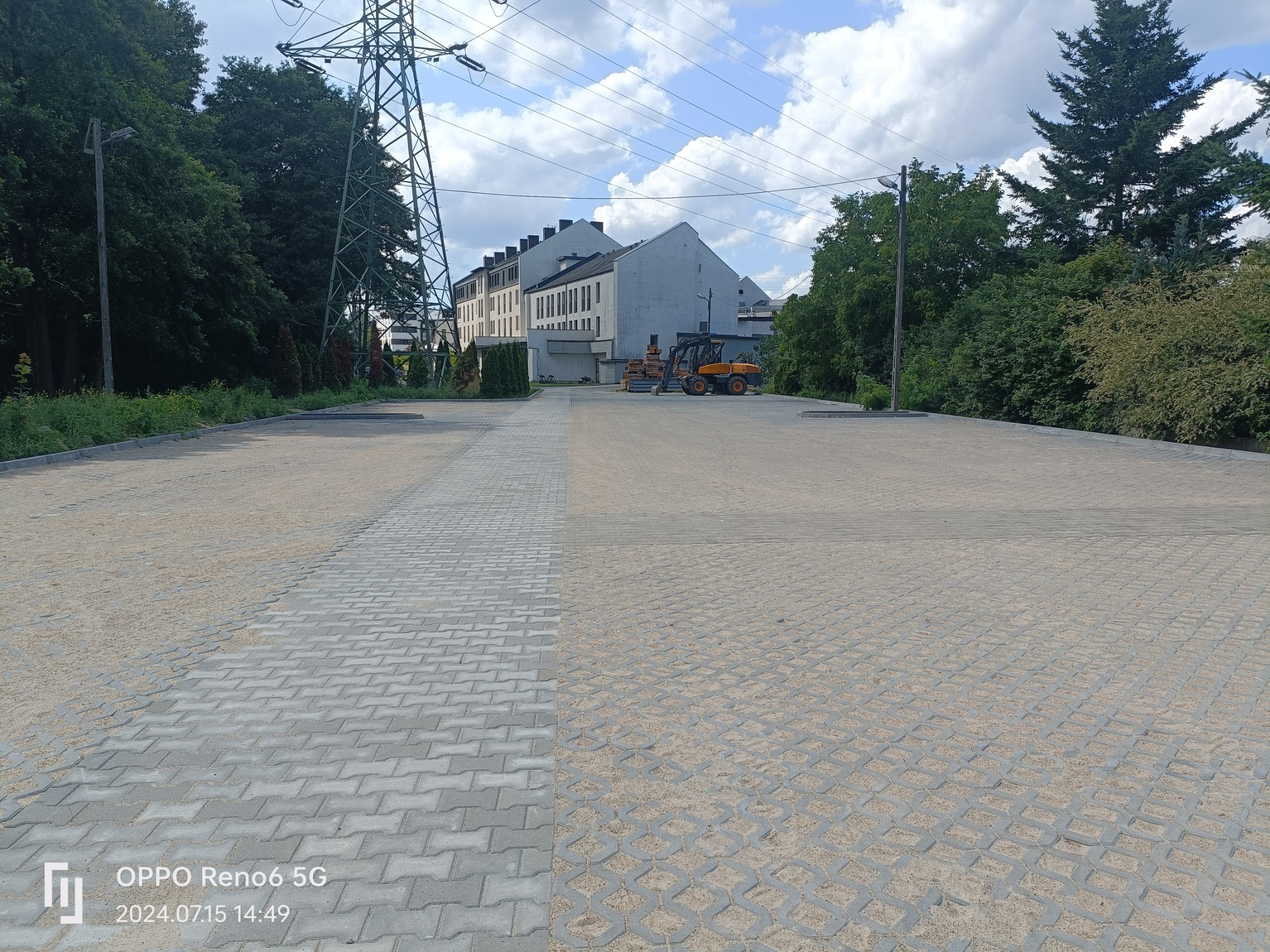 Nowo ułożona, szara kostka brukowa na dużej powierzchni parkingu lub placu, z widocznymi śladami piasku w spoinach, z maszyną budowlaną w tle.