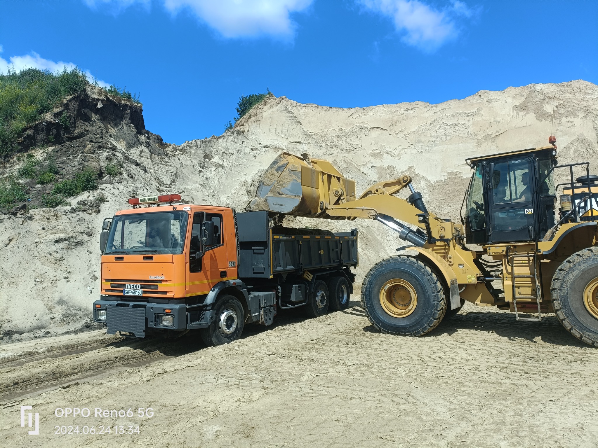 Żółta ładowarka CAT nabiera piasek z hałdy i wsypuje do pomarańczowej ciężarówki Iveco z napisem Służba Drogowa, w tle widać dużą górę piasku i błękitne niebo.