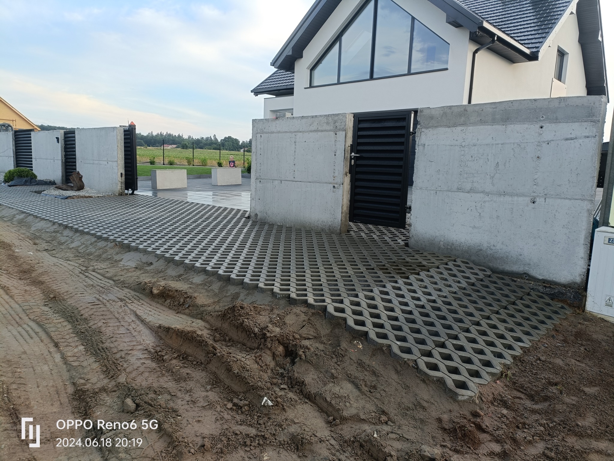 Ułożona ażurowa nawierzchnia z betonowych płyt na podjeździe przed nowoczesnym domem z betonowymi słupkami ogrodzenia i czarną bramą wjazdową, częściowo zanieczyszczona błotem.