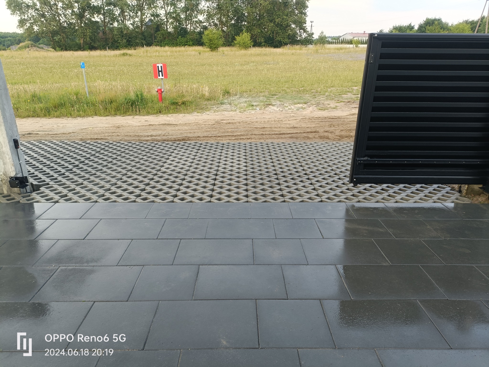 Ułożona, mokra, szara kostka brukowa przed domem, z widoczną trawnikową kratką parkingową i czarną bramą w tle.