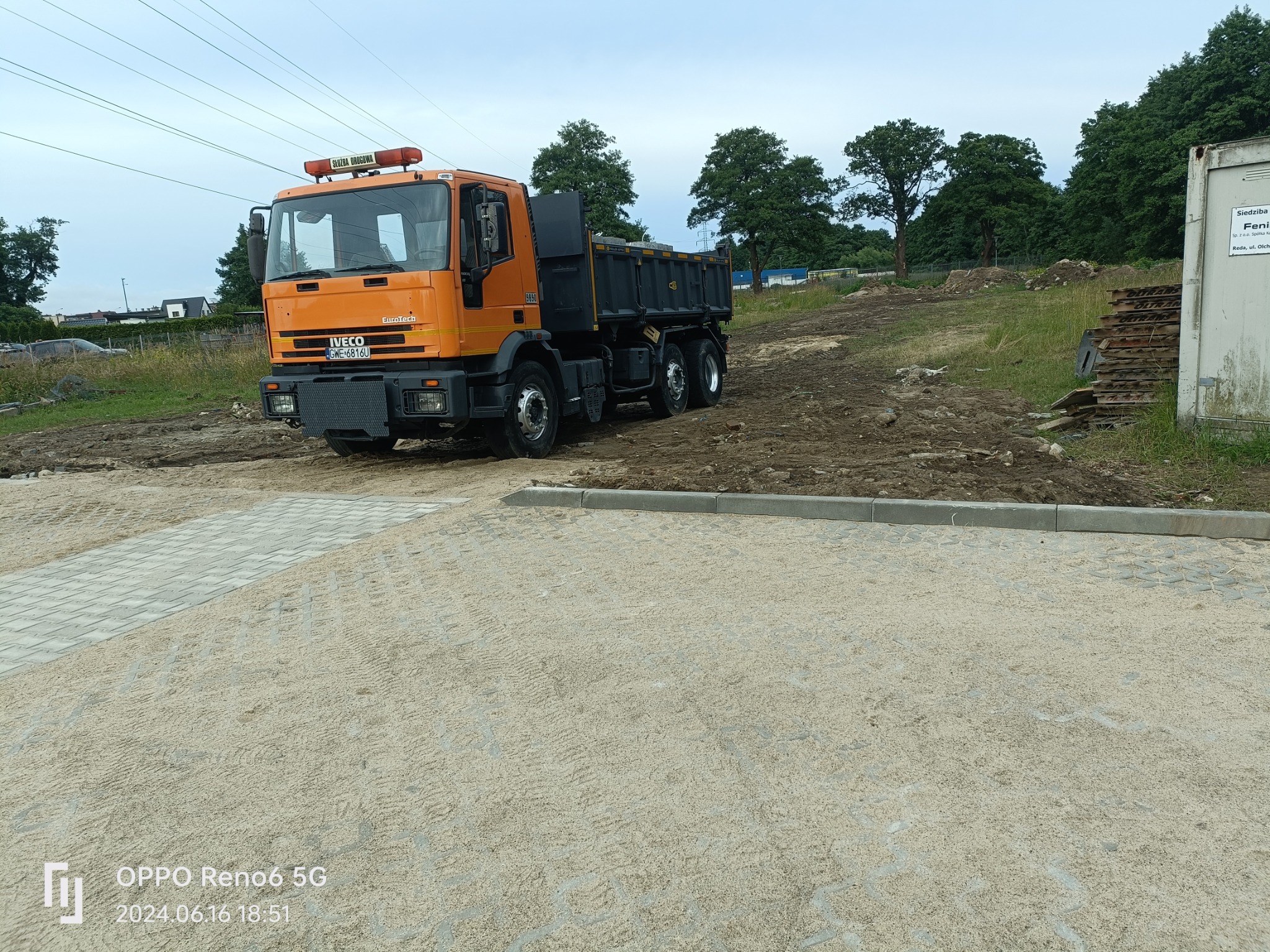 Pomarańczowy samochód ciężarowy Iveco EuroTech na placu budowy z częściowo ułożoną nawierzchnią z ażurowych płyt betonowych, w tle ziemia i drzewa.