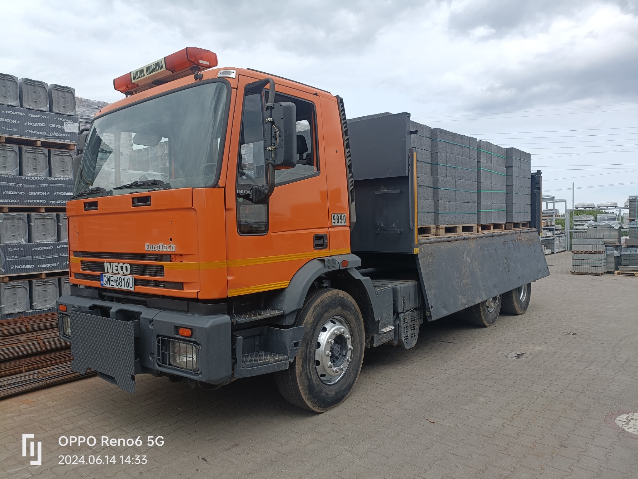 Pomarańczowy samochód ciężarowy Iveco z napisem 'Służba Drogowa' na dachu, załadowany paletami szarych kostek brukowych, stojący na placu wyłożonym kostką brukową. W tle składowane palety kostki.