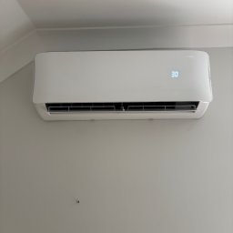 Hydrosław Sławomir Wojtalik - Biała klimatyzacja IMOTO na szarej ścianie, ustawiona na 30 stopni, widok z dołu, blisko sufitu.