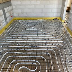 Hydrosław Sławomir Wojtalik - Białe rury ogrzewania podłogowego marki Keller, ułożone na folii z czarną siatką, przytwierdzone spinkami, w surowym wnętrzu z żółtą taśmą przy ścianach.