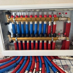 Hydrosław Sławomir Wojtalik - Rozdzielacz ogrzewania podłogowego z numerowanymi zaworami, rurami w izolacji czerwonej i niebieskiej, zamontowany w białej szafce. Widoczna folia izolacyjna na podłodze.