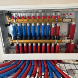 Rozdzielacz ogrzewania podłogowego z numerowanymi zaworami, rurami w izolacji czerwonej i niebieskiej, zamontowany w białej szafce. Widoczna folia izolacyjna na podłodze.