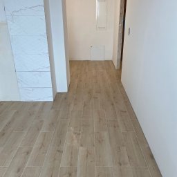 Hydrosław Sławomir Wojtalik - Ukończona podłoga z płytek imitujących drewno w jasnym odcieniu, kontrastująca z białą ścianą i fragmentem marmurowej okładziny. Widoczny fragment instalacji elektrycznej.