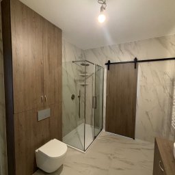 Nowoczesna łazienka z kabiną prysznicową, podwieszaną toaletą i przesuwnymi drzwiami w stylu stodoły. Marmurowe płytki na ścianach i podłodze, drewniane szafki.