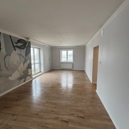 Jasne wnętrze po remoncie: drewniana podłoga, białe ściany z dekoracyjnym muralem kwiatowym, okna z widokiem na zewnątrz. Minimalistyczny design, świeżo po wykończeniu.