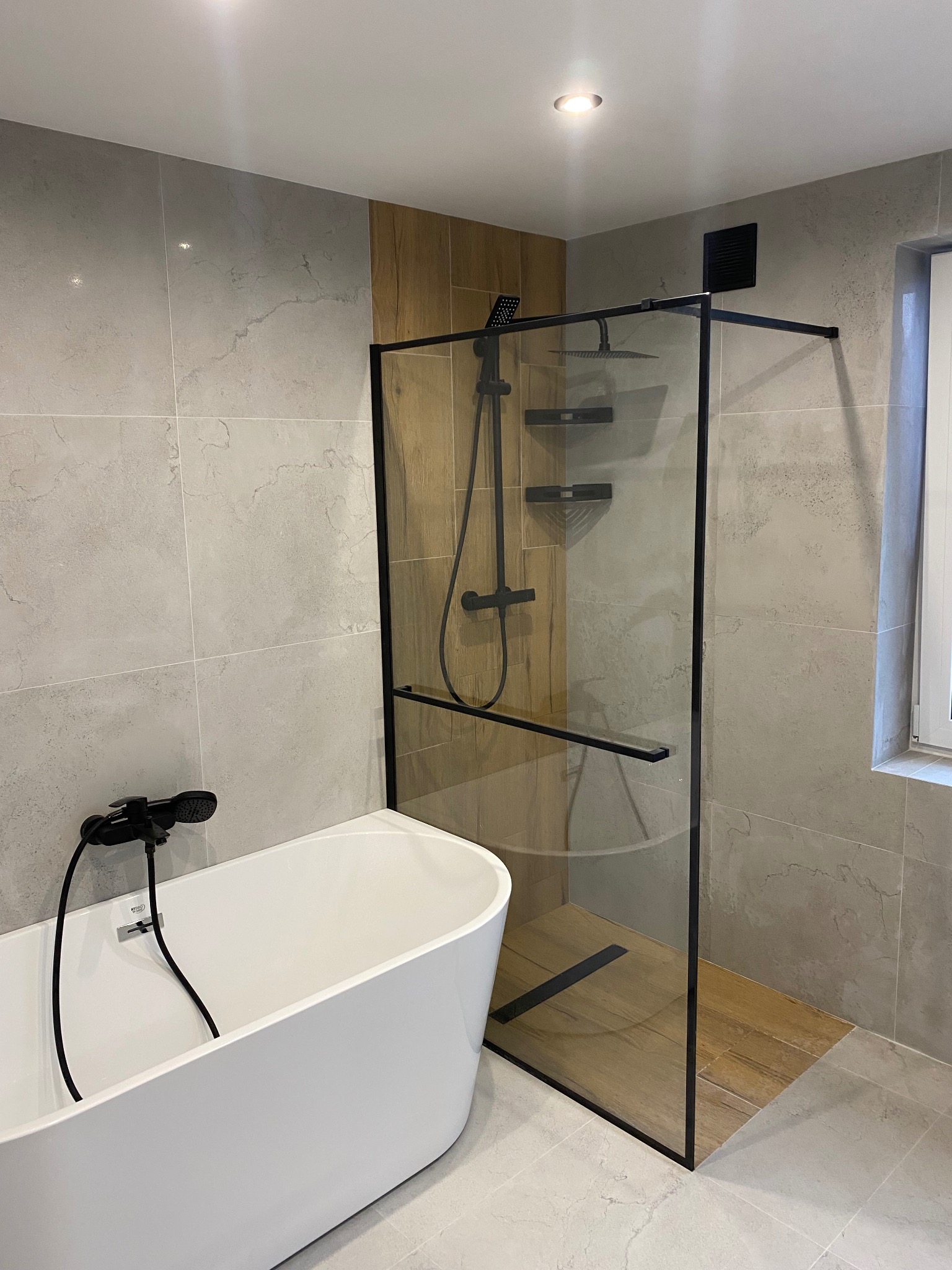 Nowoczesna łazienka z wolnostojącą wanną i kabiną prysznicową typu walk-in z czarnymi profilami. Szare płytki imitujące marmur i drewniane akcenty.