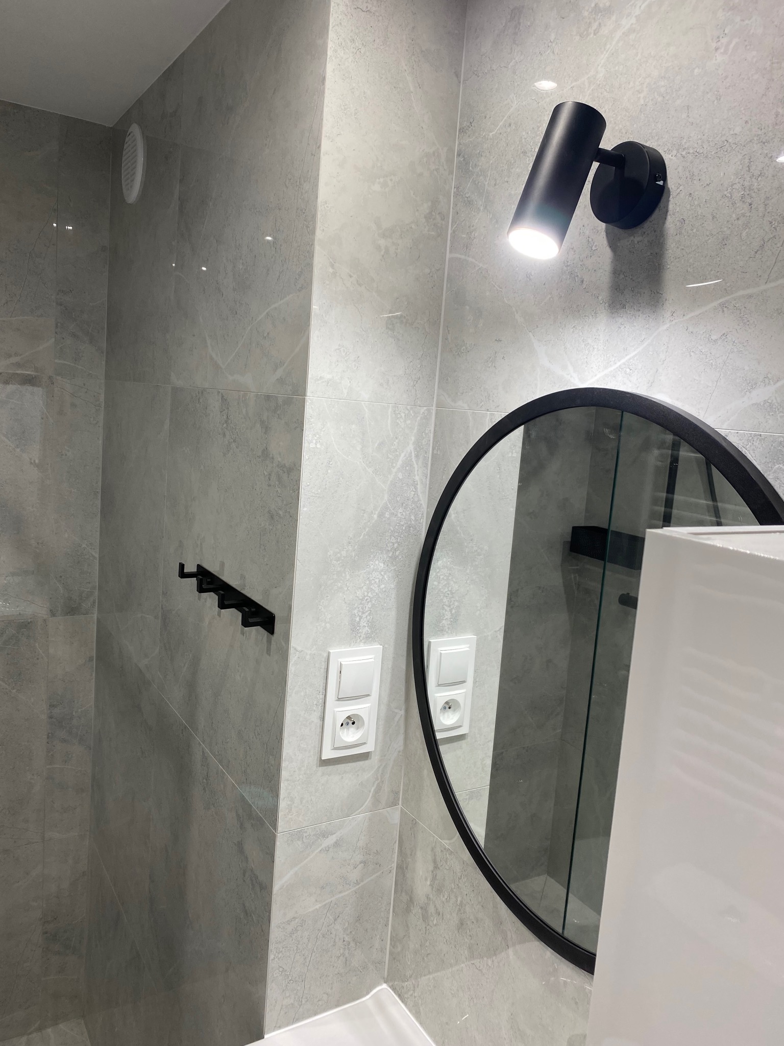Wykończona łazienka z szarymi płytkami imitującymi marmur, okrągłym lustrem i czarnymi akcentami: lampą i wieszakiem na ręczniki. Widoczny fragment białej umywalki.