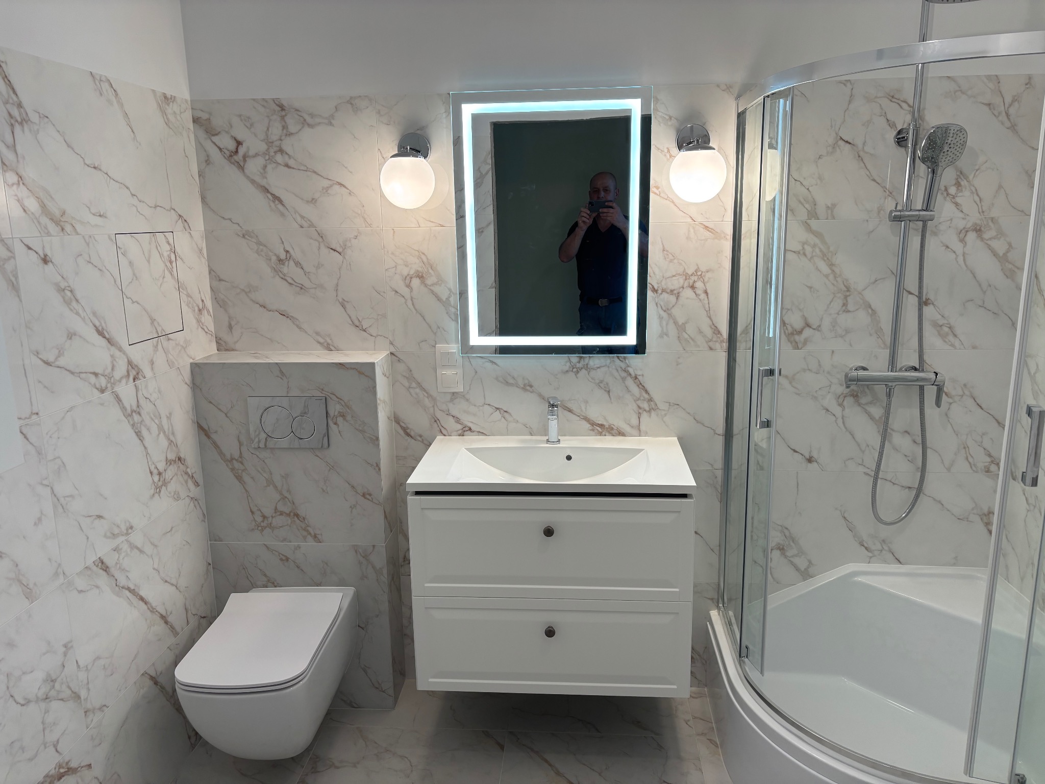 Wykończona łazienka z kabiną prysznicową, umywalką z szafką i podwieszanym WC, marmurowe płytki, oświetlone lustro z odbiciem fotografa.
