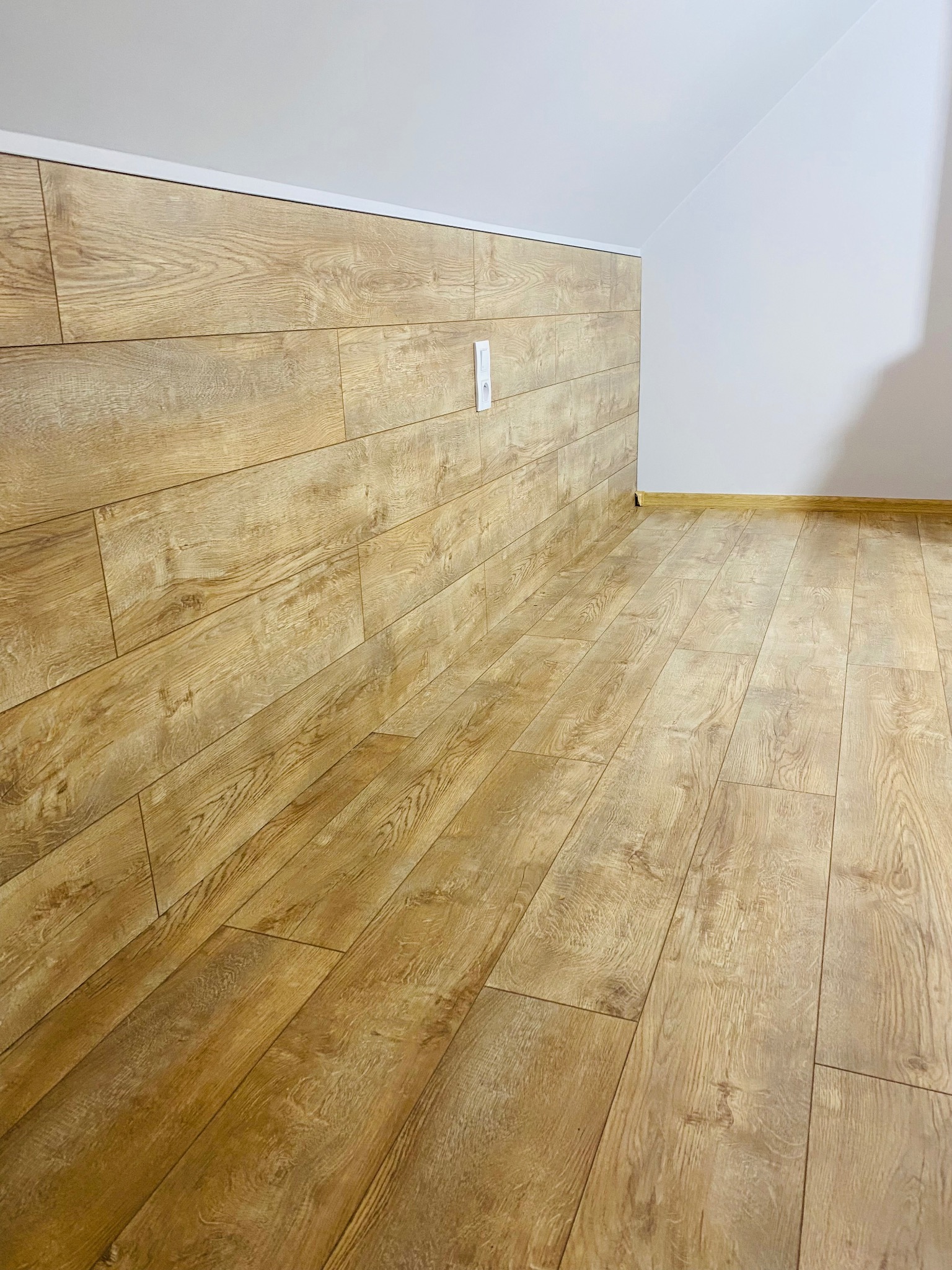 Jasne panele podłogowe i ścienne imitujące drewno w nowym pokoju na poddaszu, z widocznym gniazdkiem elektrycznym na ścianie.