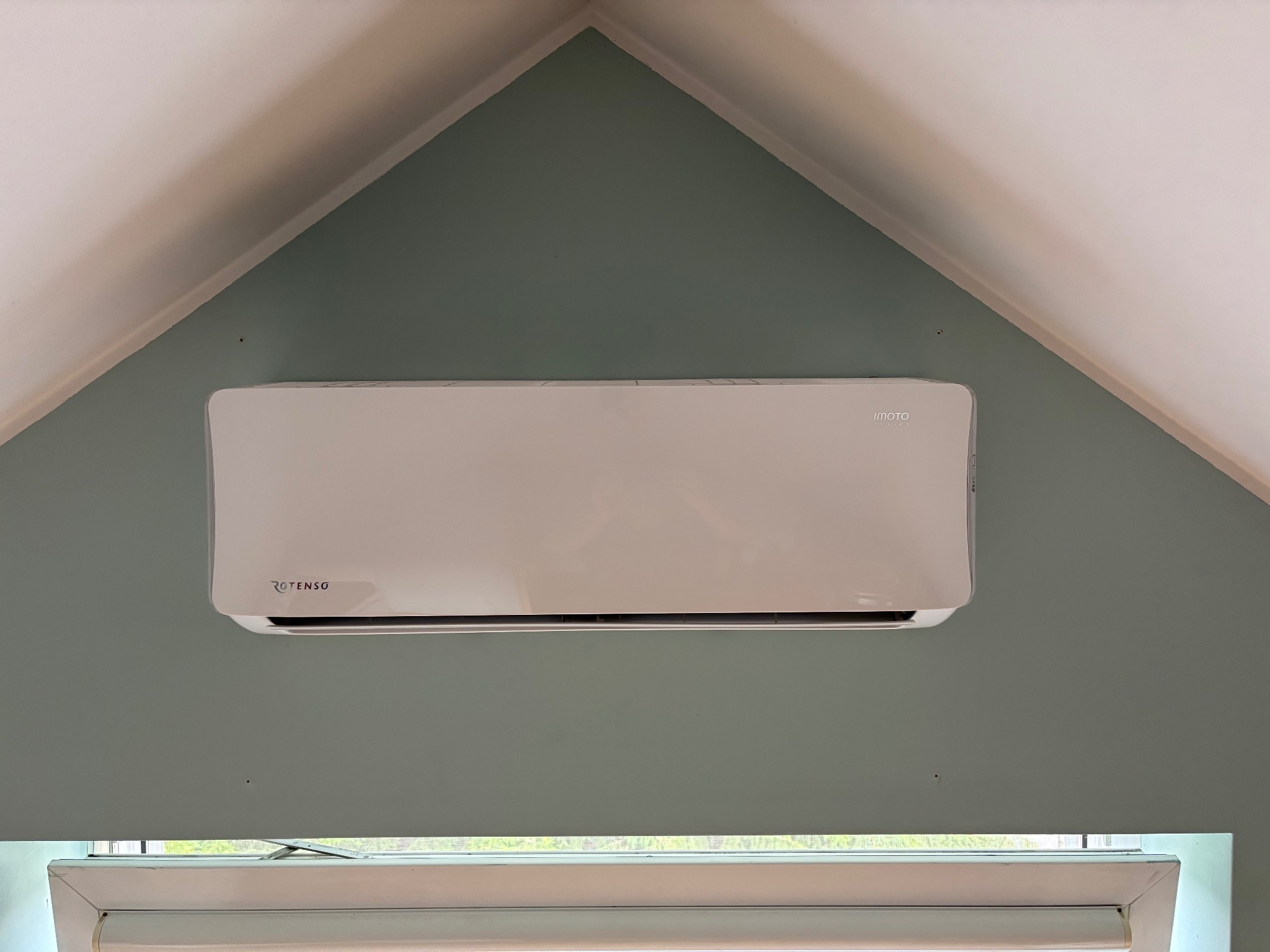 Biały klimatyzator Rotenso zamontowany na zielonej ścianie pod skośnym sufitem, widok z dołu, okno w tle.